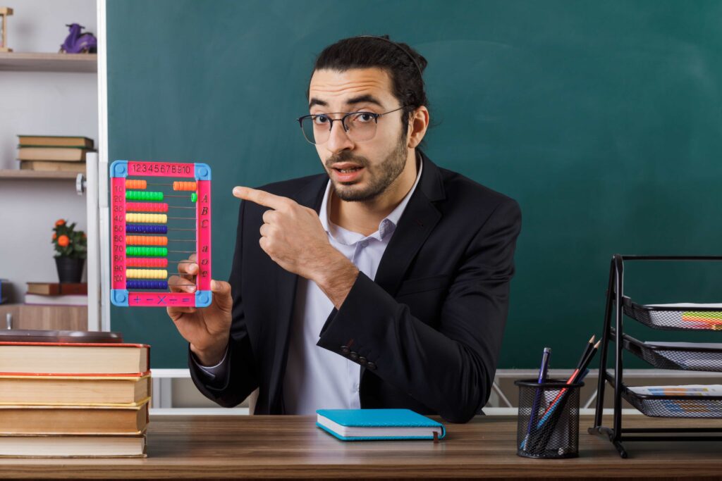 surprised male teacher wearing glasses holding points abacus sitting table with school tools classroom 1